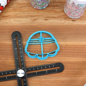 May include: A blue plastic cookie cutter shaped like a flying saucer with two small circles on top. The cutter is on a wooden surface with a ruler and other baking supplies.