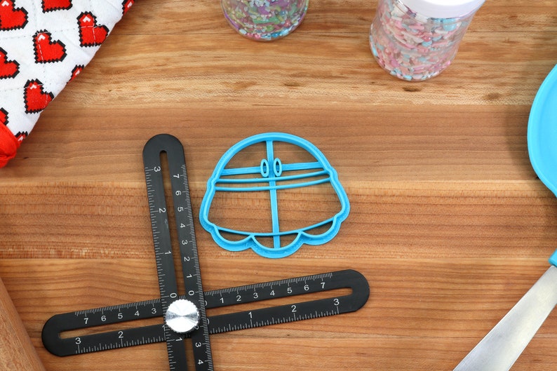 May include: A blue plastic cookie cutter shaped like a flying saucer with two small circles on top. The cutter is on a wooden surface with a ruler and other baking supplies.
