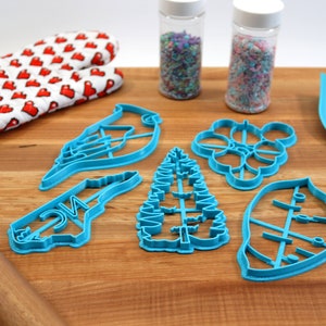 May include: A set of six blue plastic cookie cutters shaped like a bird, a tree, a leaf, a flower, and the state of North Carolina. The cutters are on a wooden cutting board with a rolling pin, a spatula, and two jars of sprinkles.