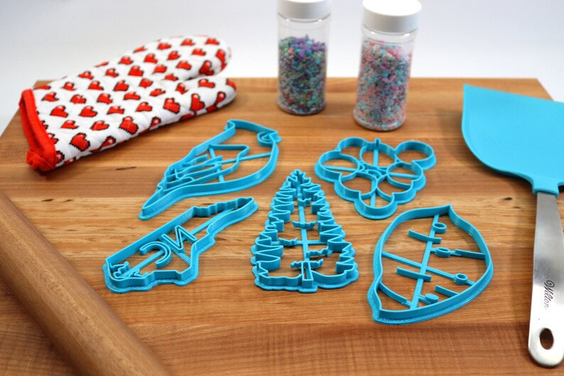 May include: A set of six blue plastic cookie cutters shaped like a bird, a tree, a leaf, a flower, and the state of North Carolina. The cutters are on a wooden cutting board with a rolling pin, a spatula, and two jars of sprinkles.