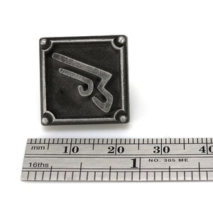 May include: A silver-colored metal pin with a stylized symbol on a black background. The pin is approximately 2 cm wide.