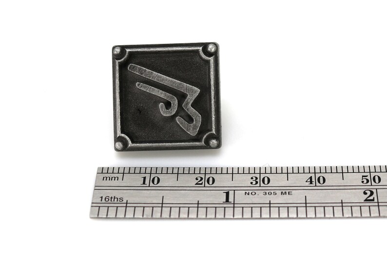 May include: A silver-colored metal pin with a stylized symbol on a black background. The pin is approximately 2 cm wide.
