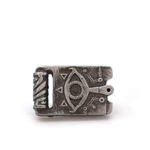 May include: A silver metal charm with a detailed design of an eye with a symbol above it. The charm has a textured surface and a small hole at the top.