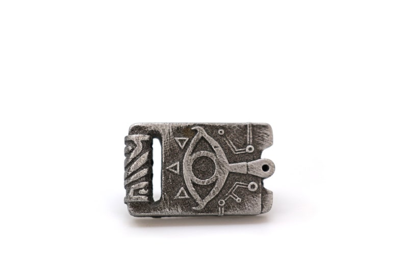 May include: A silver metal charm with a detailed design of an eye with a symbol above it. The charm has a textured surface and a small hole at the top.