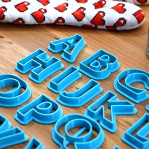 May include: A set of blue plastic alphabet cookie cutters on a wooden surface. The cutters are arranged in alphabetical order.