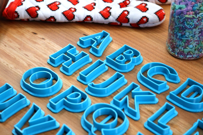 May include: A set of blue plastic alphabet cookie cutters on a wooden surface. The cutters are arranged in alphabetical order.