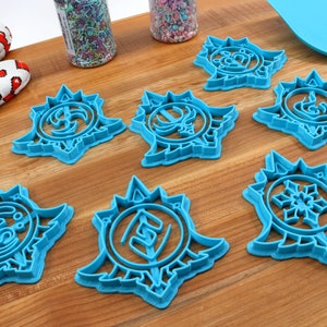 May include: Seven blue plastic cookie cutters shaped like snowflakes with different designs in the center. The cutters are on a wooden surface.