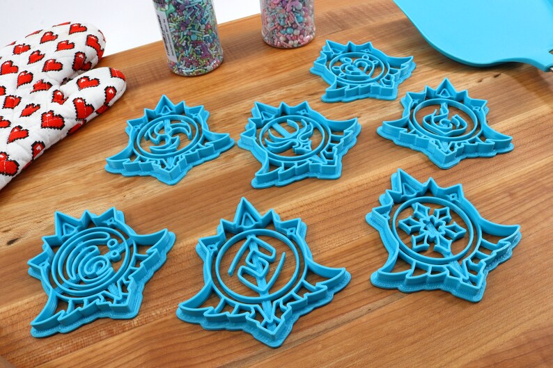 May include: Seven blue plastic cookie cutters shaped like snowflakes with different designs in the center. The cutters are on a wooden surface.