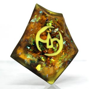 May include: A yellow and green iridescent resin dice with a yellow symbol of a stylized "5" on one face.
