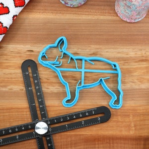Puede incluir: Un cortador de galletas de pl&aacute;stico azul con forma de Bulldog Franc&eacute;s. El cortador est&aacute; sobre una superficie de madera con una regla negra y una tela blanca y roja con corazones.