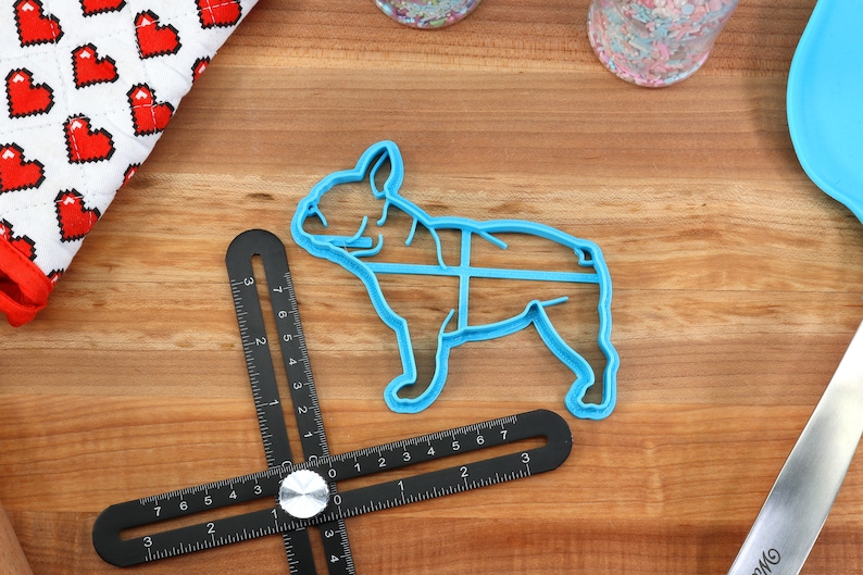 Puede incluir: Un cortador de galletas de pl&aacute;stico azul con forma de Bulldog Franc&eacute;s. El cortador est&aacute; sobre una superficie de madera con una regla negra y una tela blanca y roja con corazones.