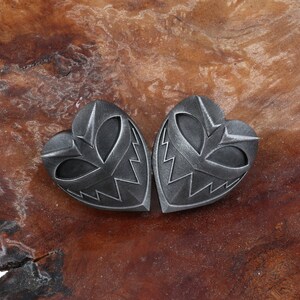 May include: Two silver heart-shaped pendants with a detailed, menacing face design. The pendants are on a brown wooden surface.