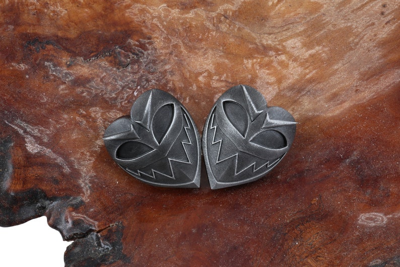 May include: Two silver heart-shaped pendants with a detailed, menacing face design. The pendants are on a brown wooden surface.