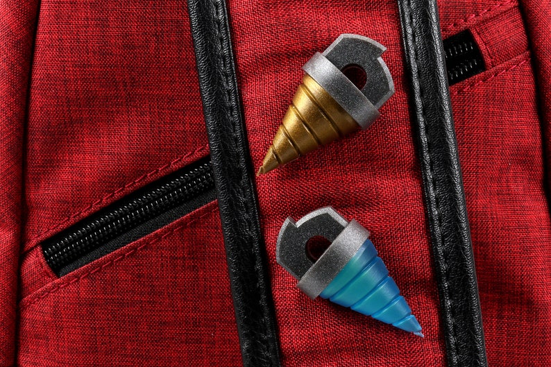 May include: Two miniature drill-shaped accessories, one gold and one blue, with silver accents, rest on a textured red fabric. The accessories have a black top with a hole. A black zipper and trim are visible.