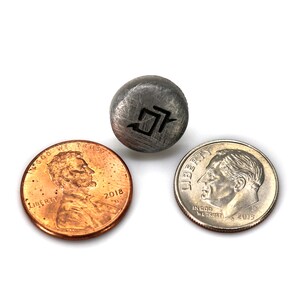 May include: A small, round, gray metal object with a black graphic is positioned between a copper penny and a silver dime. The penny is dated 2018. The dime is dated 2015.