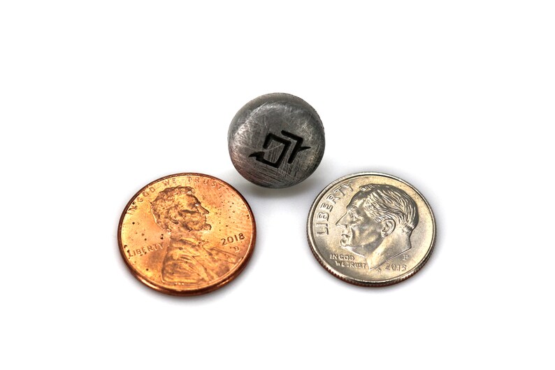 May include: A small, round, gray metal object with a black graphic is positioned between a copper penny and a silver dime. The penny is dated 2018. The dime is dated 2015.