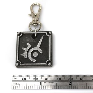 May include: A silver metal keychain with a black square charm featuring a stylized silver design. The charm has a silver ring attached to a silver metal split ring.
