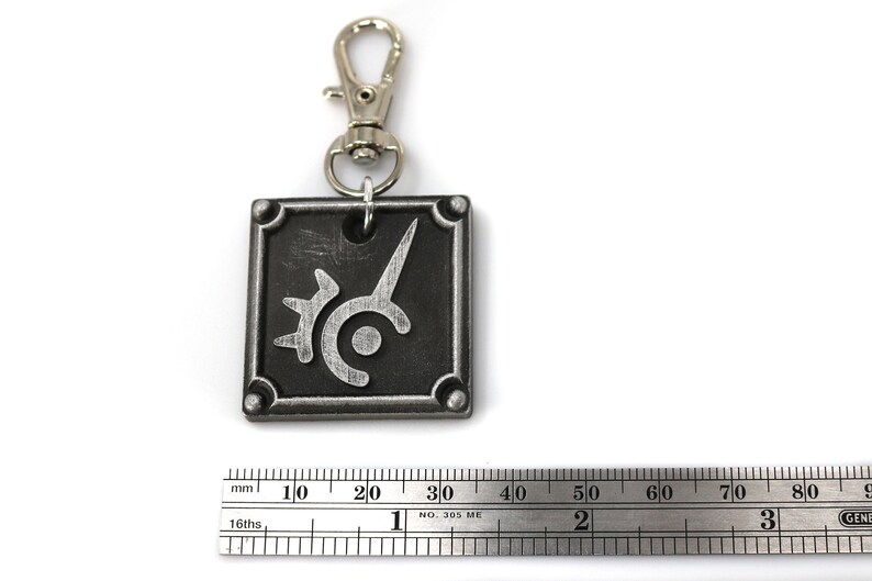 May include: A silver metal keychain with a black square charm featuring a stylized silver design. The charm has a silver ring attached to a silver metal split ring.
