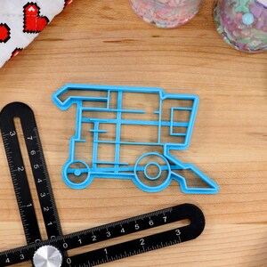 May include: A blue plastic cookie cutter shaped like a combine harvester. The cutter is on a wooden surface with a ruler and other baking supplies.