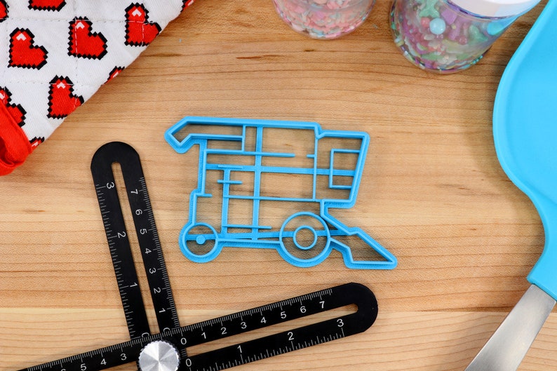 May include: A blue plastic cookie cutter shaped like a combine harvester. The cutter is on a wooden surface with a ruler and other baking supplies.