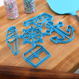 May include: A collection of blue plastic nautical-themed cookie cutters, including an anchor, ship's wheel, and sailboat, arranged on a wooden cutting board. Also present are a blue spatula, a heart-patterned oven mitt, and jars of sprinkles.