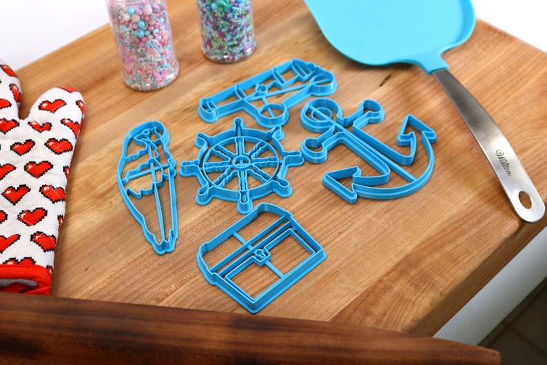 May include: A collection of blue plastic nautical-themed cookie cutters, including an anchor, ship's wheel, and sailboat, arranged on a wooden cutting board. Also present are a blue spatula, a heart-patterned oven mitt, and jars of sprinkles.