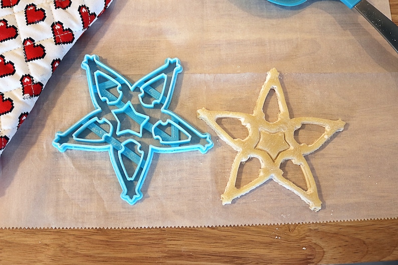 Pu&ograve; includere: Un tagliabiscotti a forma di stella azzurra accanto a un biscotto a forma di stella su carta da forno. Il tagliabiscotti ha un design dettagliato. Sullo sfondo, una presina a forma di cuore rosso e bianco.