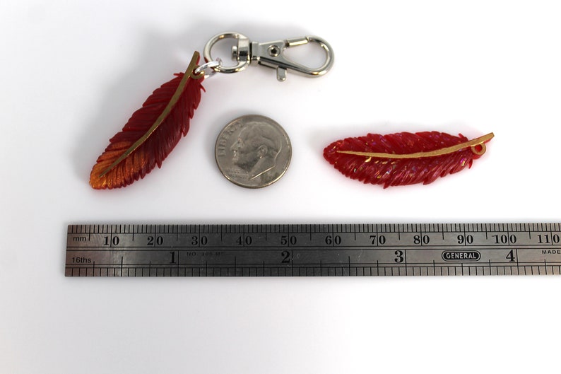 May include: Two red and gold feather charms, one with a silver key ring. The charms are approximately 2 inches long and are next to a US dime for size comparison.