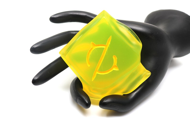 May include: A yellow and green soap bar with a stylized symbol on the front. The soap is being held in a black hand.