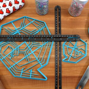 May include: Two blue plastic snowflake-shaped cookie cutters on a wooden surface with a black ruler, a rolling pin, a blue spatula, and two jars of sprinkles.