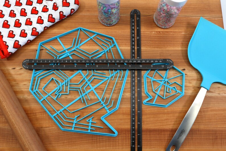 May include: Two blue plastic snowflake-shaped cookie cutters on a wooden surface with a black ruler, a rolling pin, a blue spatula, and two jars of sprinkles.