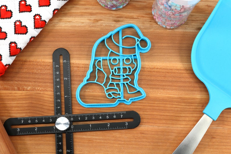May include: Blue plastic cookie cutter shaped like a snowman wearing a Santa hat. The snowman is holding a round object in its left hand.