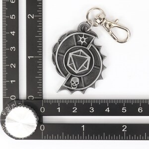 May include: A silver-toned metal keychain featuring a stylized skull and a 20-sided die. The keychain has a silver-toned metal ring and a silver-toned metal clasp.