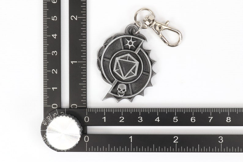 May include: A silver-toned metal keychain featuring a stylized skull and a 20-sided die. The keychain has a silver-toned metal ring and a silver-toned metal clasp.