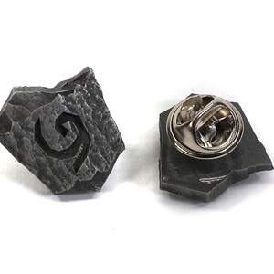 Puede incluir: Un pin de metal gris con forma de roca con un dise&ntilde;o de espiral estilizado. La parte posterior del pin tiene un cierre de metal.