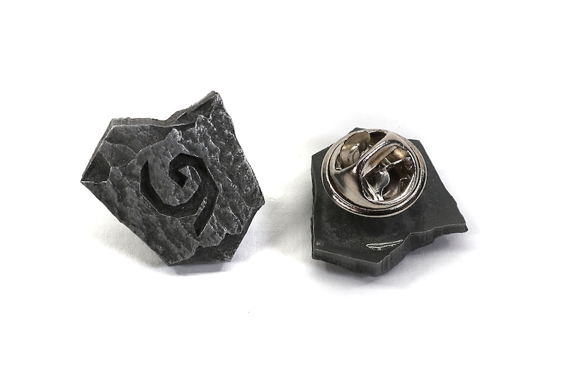 Puede incluir: Un pin de metal gris con forma de roca con un dise&ntilde;o de espiral estilizado. La parte posterior del pin tiene un cierre de metal.