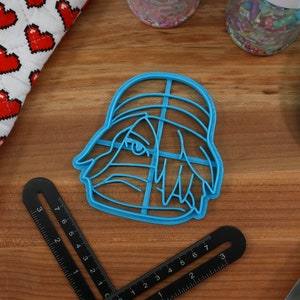 May include: A blue plastic cookie cutter shaped like a cartoon character with a hat and hair. The character has a large eye and a small mouth.