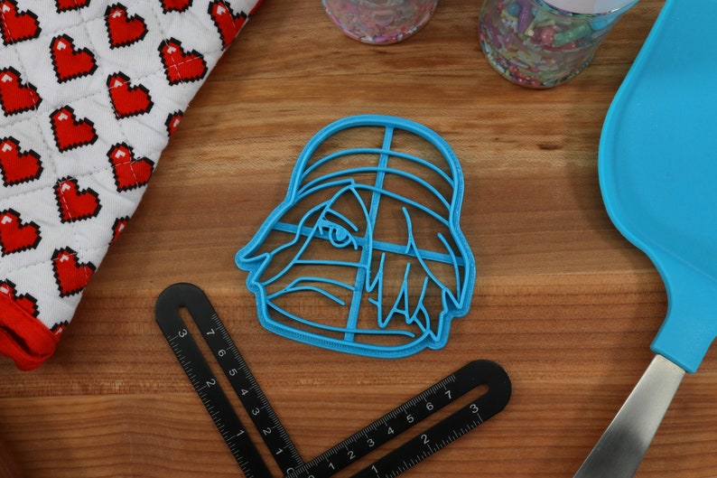 May include: A blue plastic cookie cutter shaped like a cartoon character with a hat and hair. The character has a large eye and a small mouth.