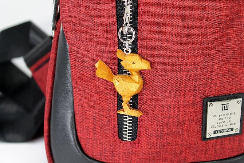 May include: A red zippered bag with a gold-colored bird charm attached to the zipper. The bag has a black leather trim and a black strap. The bag has a small black patch with the text "Where is the heart to Receive Goods where TUOWAN" on it.