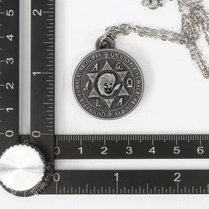 May include: A silver-colored pendant necklace with a circular pendant featuring engraved text and a central star design. The pendant is attached to a delicate chain. The image includes a ruler for scale.