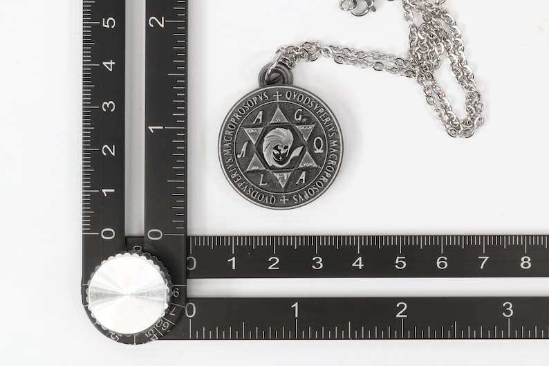 May include: A silver-colored pendant necklace with a circular pendant featuring engraved text and a central star design. The pendant is attached to a delicate chain. The image includes a ruler for scale.