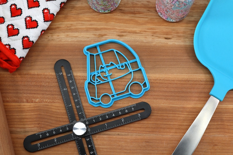 Puede incluir: Un cortador de galletas de pl&aacute;stico azul con forma de coche con una persona dentro. El cortador est&aacute; sobre una superficie de madera con una regla negra y una esp&aacute;tula azul.