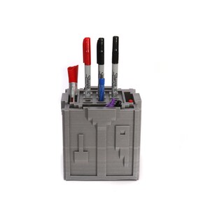 May include: Gray 3D printed pen holder with a maze design. The holder has four slots for pens and markers. There are three black Sharpie markers and one red marker in the holder.