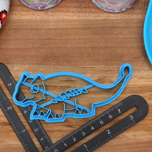 May include: A blue plastic cookie cutter shaped like a dinosaur. The cutter is on a wooden surface with a ruler and a blue silicone mat.