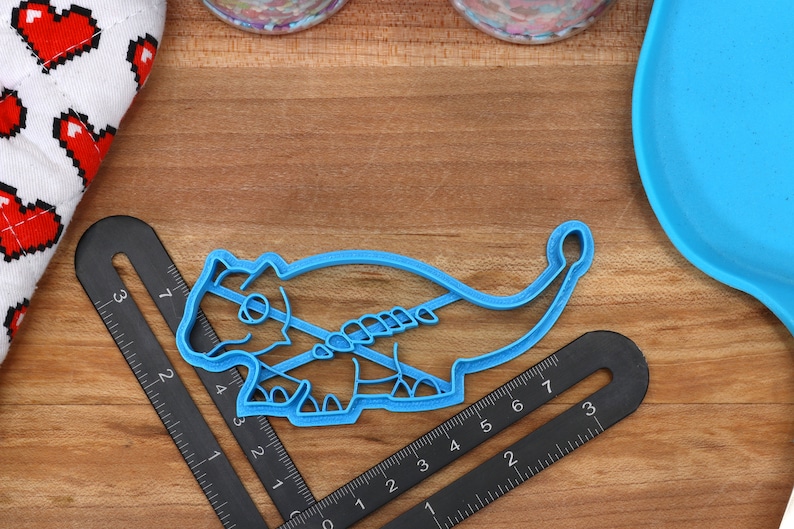 May include: A blue plastic cookie cutter shaped like a dinosaur. The cutter is on a wooden surface with a ruler and a blue silicone mat.
