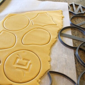 May include: A sheet of cookie dough with various shapes cut out using cookie cutters. The cookie cutters are shown on the right side of the image. The cookie cutters are gray and made of plastic.