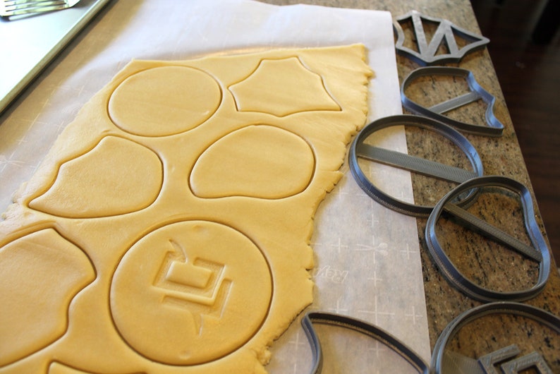 May include: A sheet of cookie dough with various shapes cut out using cookie cutters. The cookie cutters are shown on the right side of the image. The cookie cutters are gray and made of plastic.