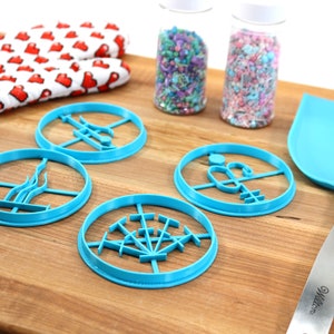 May include: Four blue plastic cookie cutters shaped like different designs on a wooden chopping board. There are two jars of sprinkles in the background.