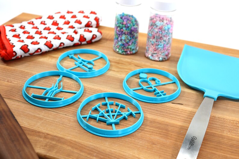 May include: Four blue plastic cookie cutters shaped like different designs on a wooden chopping board. There are two jars of sprinkles in the background.