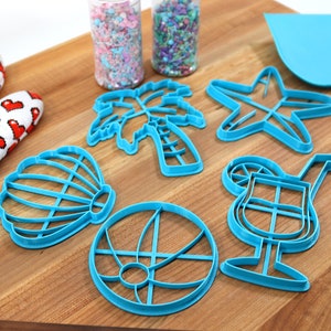 May include: A set of blue plastic cookie cutters shaped like a palm tree, seashell, starfish, beach ball, and cocktail glass. The cutters are on a wooden cutting board with two jars of sprinkles in the background.